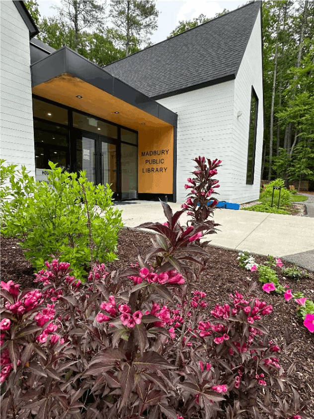 Modern white brick library building with a black roof, large windows, and a sign reading Madbury Public Library. Pink flowering plants and green shrubs are in the foreground, surrounded by trees.